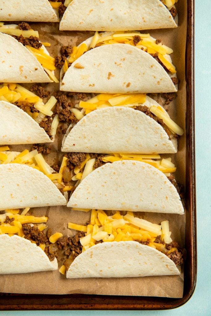 Sheet Pan Tacos are the easiest way to get crispy, cheesy, restaurant-worthy baked tacos on the table ready to serve all at the same time! A baking sheet lined with parchment paper holds several soft tacos filled with seasoned taco meat and shredded yellow and white cheese. The tacos are arranged neatly in rows on a sheet pan.