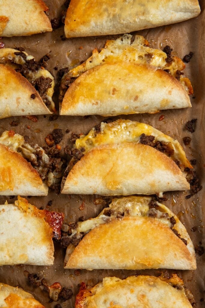 Sheet Pan Tacos are the easiest way to get crispy, cheesy, restaurant-worthy baked tacos on the table ready to serve all at the same time! A baking sheet lined with parchment paper holds several crispy baked tacos filled with seasoned ground beef and melted cheese, arranged in neat rows.