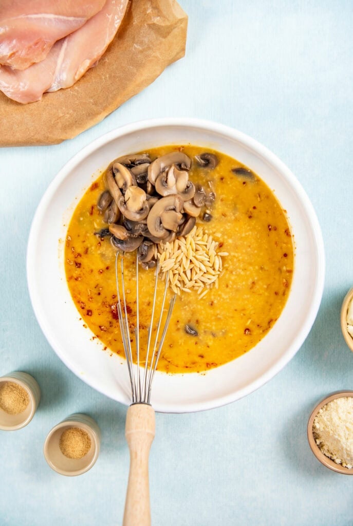 This dump-and-bake Chicken Orzo Bake is creamy and cheesy with serious flavor in every bite. One dish, simple ingredients, huge flavor! A white bowl contains a yellow-orange sauce with sliced mushrooms and orzo pasta being mixed with a whisk. Surrounding the bowl are small bowls of seasonings and raw chicken on parchment paper.