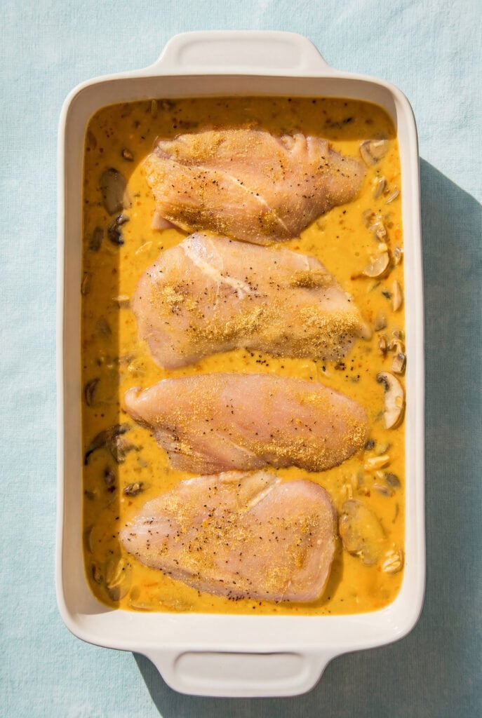 This dump-and-bake Chicken Orzo Bake is creamy and cheesy with serious flavor in every bite. One dish, simple ingredients, huge flavor! Raw chicken breasts and orzo pasta sit in a rectangular baking dish filled with a creamy orange sauce and mushrooms, ready to be cooked. The dish rests on a light blue surface.