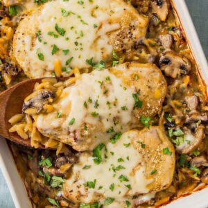 A baked casserole dish with three cheesy chicken breasts on top of a bed of rice, mushrooms, and vegetables, garnished with chopped parsley. A wooden spoon is lifting one piece of chicken.