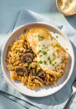 A plate with creamy chicken orzo bake with mushrooms on a white plate garnished with chopped parsley. A small bowl of grated parmesan cheese is in the background.