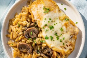 A plate with creamy chicken orzo bake with mushrooms on a white plate garnished with chopped parsley. A small bowl of grated parmesan cheese is in the background.