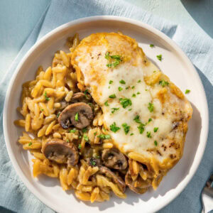 A plate with creamy chicken orzo bake with mushrooms on a white plate garnished with chopped parsley. A small bowl of grated parmesan cheese is in the background.