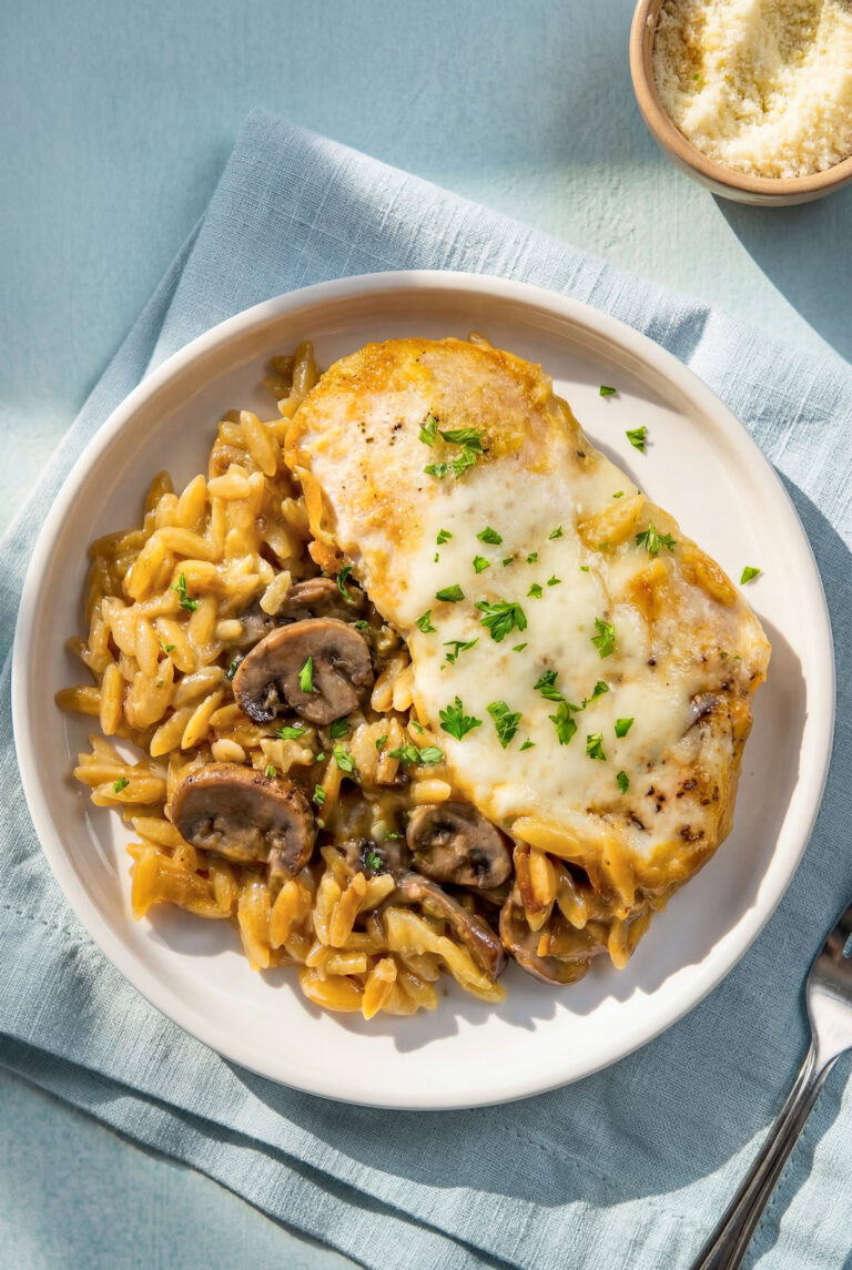 A plate with creamy chicken orzo bake with mushrooms on a white plate garnished with chopped parsley. A small bowl of grated parmesan cheese is in the background.