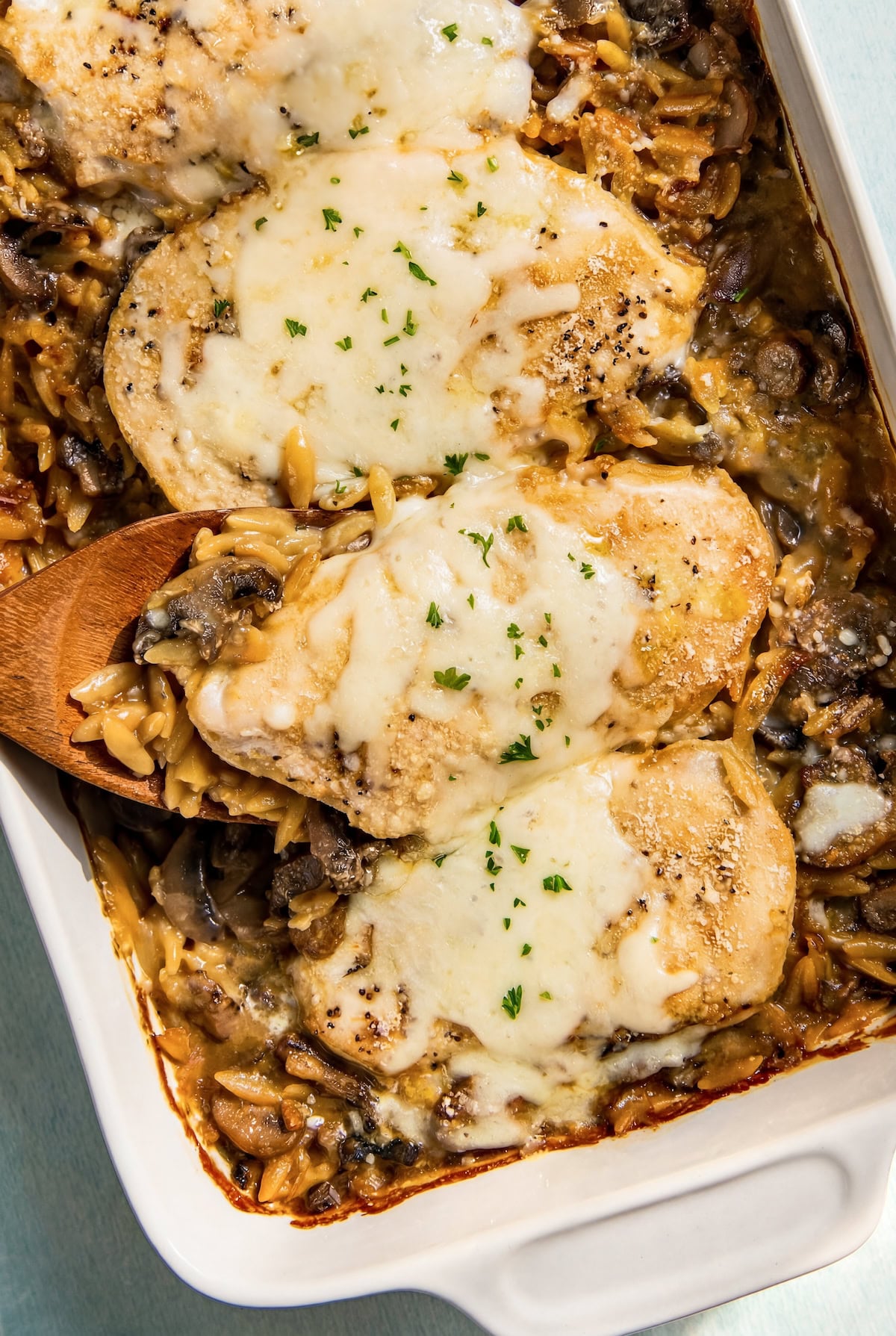 This dump-and-bake Chicken Orzo Bake is creamy and cheesy with serious flavor in every bite. One dish, simple ingredients, huge flavor! A casserole dish filled with a creamy chicken and orzo bake topped with melted cheese and fresh herbs. A wooden spoon is lifting one piece of chicken from the dish.