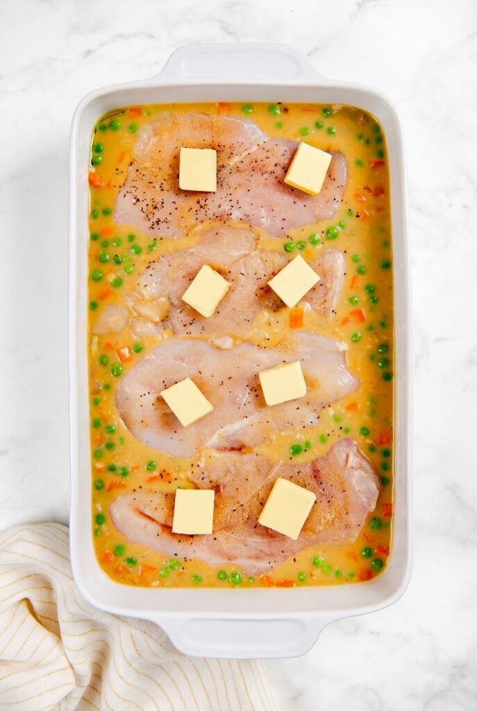 This easy chicken and rice casserole recipe is pure creamy, cheesy comfort food every single time. One pan, 5 minutes of prep, and dinner is done! Raw chicken breasts in a white casserole dish with creamy sauce, peas, diced carrots, and slices of butter on top, ready to be baked.