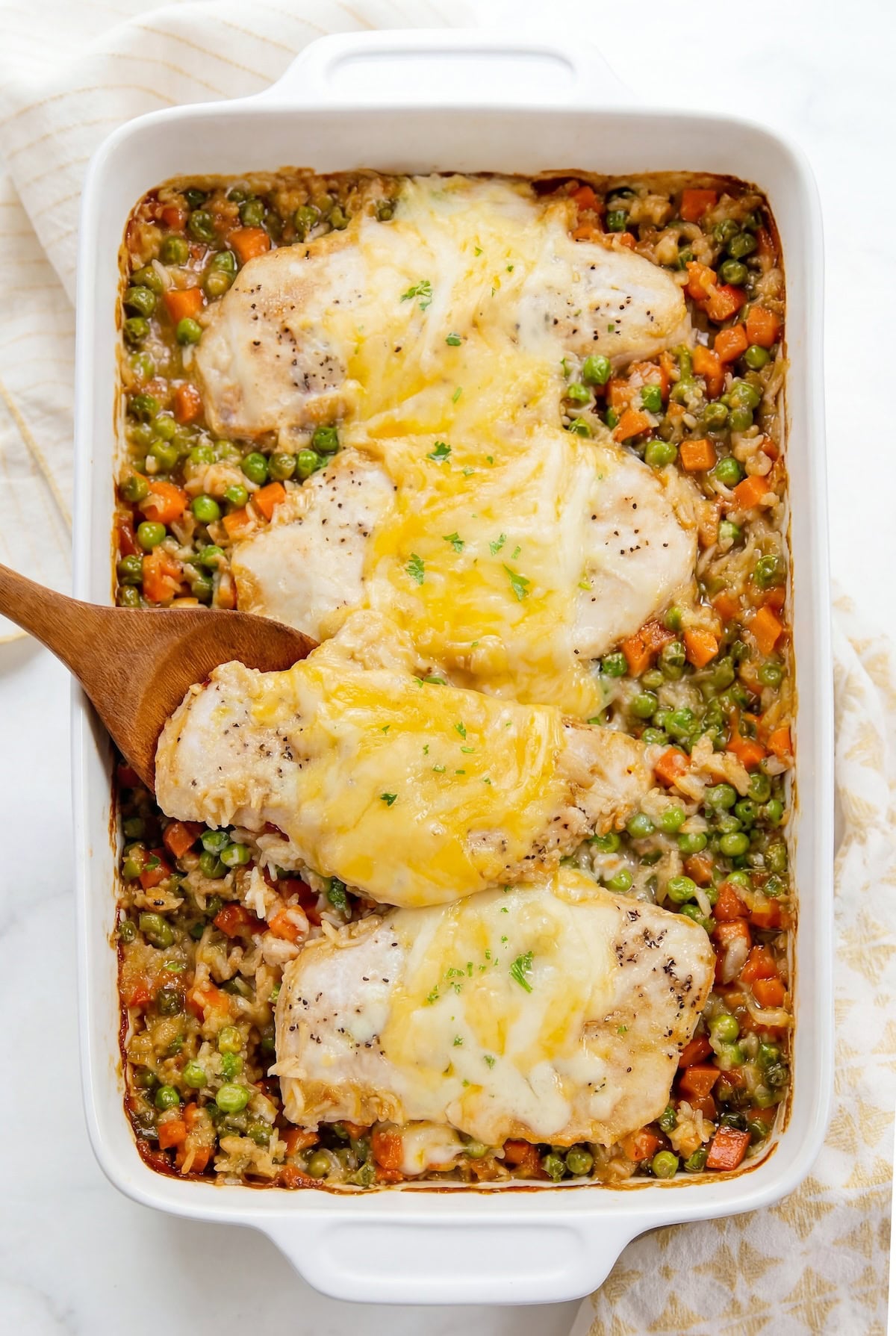 This easy chicken and rice casserole recipe is pure creamy, cheesy comfort food every single time. One pan, 5 minutes of prep, and dinner is done! A white baking dish filled with baked chicken rice casserole with peas and carrots topped with melted cheese. A wooden spoon is lifting one piece of baked chicken topped with more melted cheese.