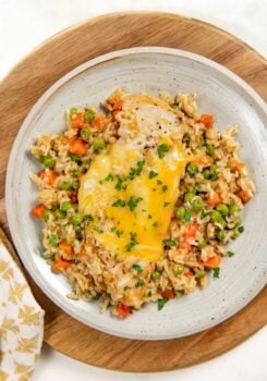 A plate of easy chicken and rice casserole with peas and diced carrots is topped with melted cheese and garnished with chopped parsley. Served on a light gray plate atop a wooden board.