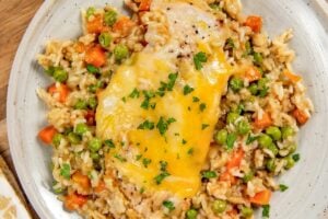 A plate of easy chicken and rice casserole with peas and diced carrots is topped with melted cheese and garnished with chopped parsley. Served on a light gray plate atop a wooden board.
