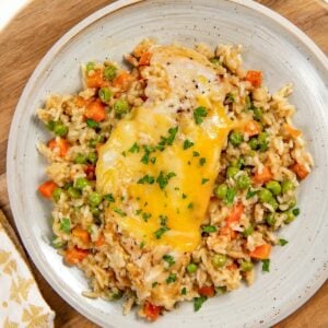 A plate of easy chicken and rice casserole with peas and diced carrots is topped with melted cheese and garnished with chopped parsley. Served on a light gray plate atop a wooden board.