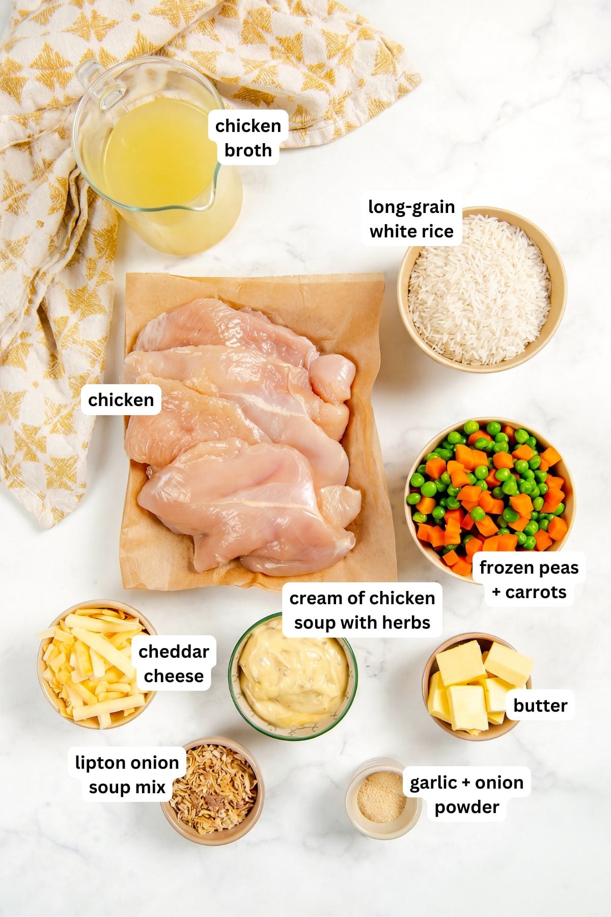 This easy chicken and rice casserole recipe is pure creamy, cheesy comfort food every single time. One pan, 5 minutes of prep, and dinner is done! Ingredients for a chicken and rice casserole recipe arranged in bowls: chicken broth, long-grain white rice, raw chicken breasts, frozen peas and carrots, cheddar cheese, cream of chicken soup with herbs, butter, Lipton onion soup mix, garlic powder, and onion powder.