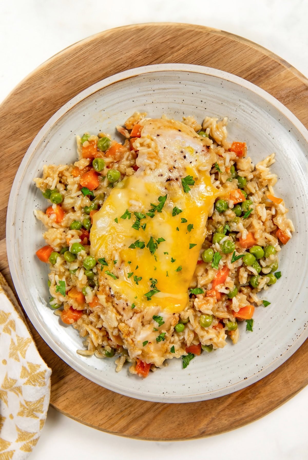 This easy chicken and rice casserole recipe is pure creamy, cheesy comfort food every single time. One pan, 5 minutes of prep, and dinner is done! A plate of easy chicken and rice casserole with peas and diced carrots is topped with melted cheese and garnished with chopped parsley. Served on a light gray plate atop a wooden board.