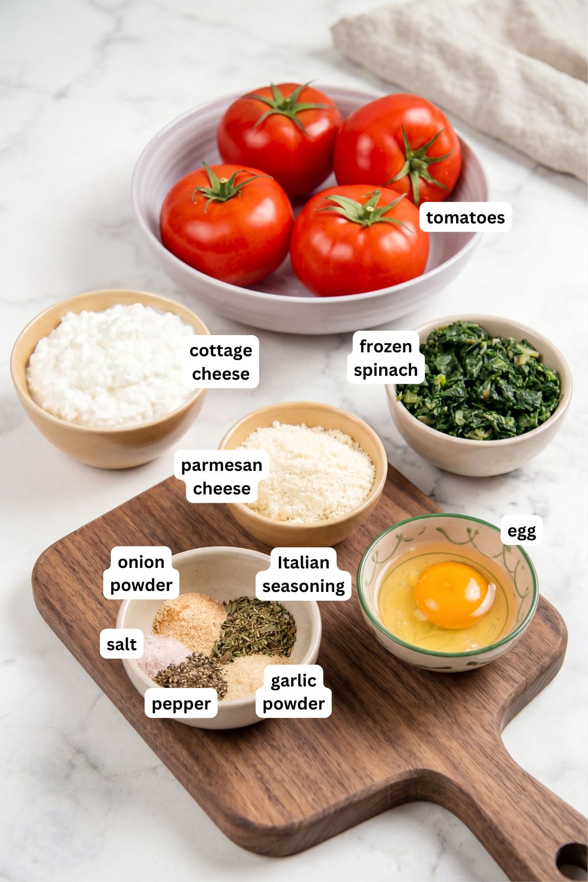 These stuffed tomatoes are ready in 30 minutes with a creamy spinach and cheese filling. Low carb, high protein, and seriously delicious! Labeled ingredients for a stuffed tomato recipe: a bowl of tomatoes, a bowl of cottage cheese, frozen spinach, a bowl of parmesan cheese, and a bowl with an egg, plus spices (salt, pepper, onion powder, garlic powder, Italian seasoning) on a wooden board.