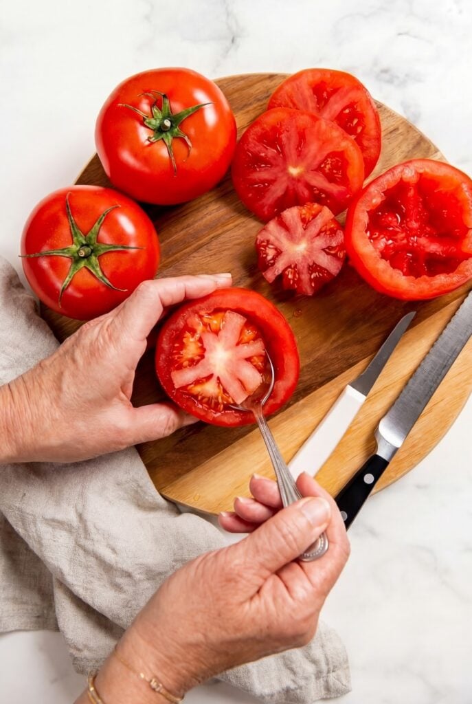 These stuffed tomatoes are ready in 30 minutes with a creamy spinach and cheese filling. Low carb, high protein, and seriously delicious! A person scoops seeds and pulp from a halved tomato with a spoon on a wooden cutting board, surrounded by whole and cut tomatoes, with a knife and a beige cloth nearby.