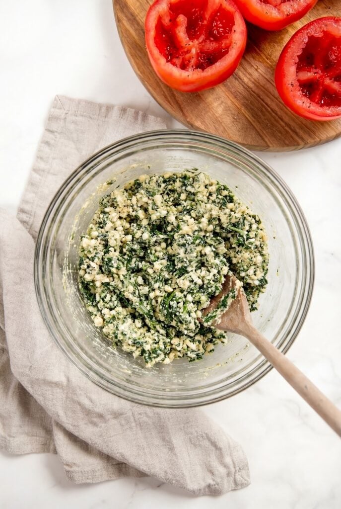 These stuffed tomatoes are ready in 30 minutes with a creamy spinach and cheese filling. Low carb, high protein, and seriously delicious! A glass bowl filled with a spinach and cheese mixture sits on a beige napkin, with a wooden spoon inside. In the background, halved and hollowed tomatoes rest on a wooden board.