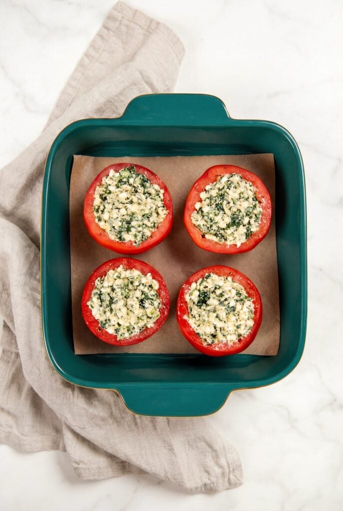 These stuffed tomatoes are ready in 30 minutes with a creamy spinach and cheese filling. Low carb, high protein, and seriously delicious! Four tomatoes are stuffed with a spinach and cheese mixture and arranged in a teal baking dish lined with parchment paper.