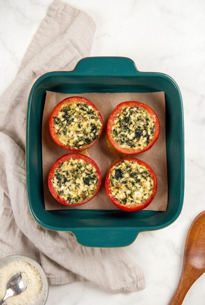 These stuffed tomatoes are ready in 30 minutes with a creamy spinach and cheese filling. Low carb, high protein, and seriously delicious! Four baked stuffed tomatoes are arranged in a square teal baking dish lined with parchment paper, next to a beige cloth, a wooden spoon, and a bowl of grated parmesan cheese.