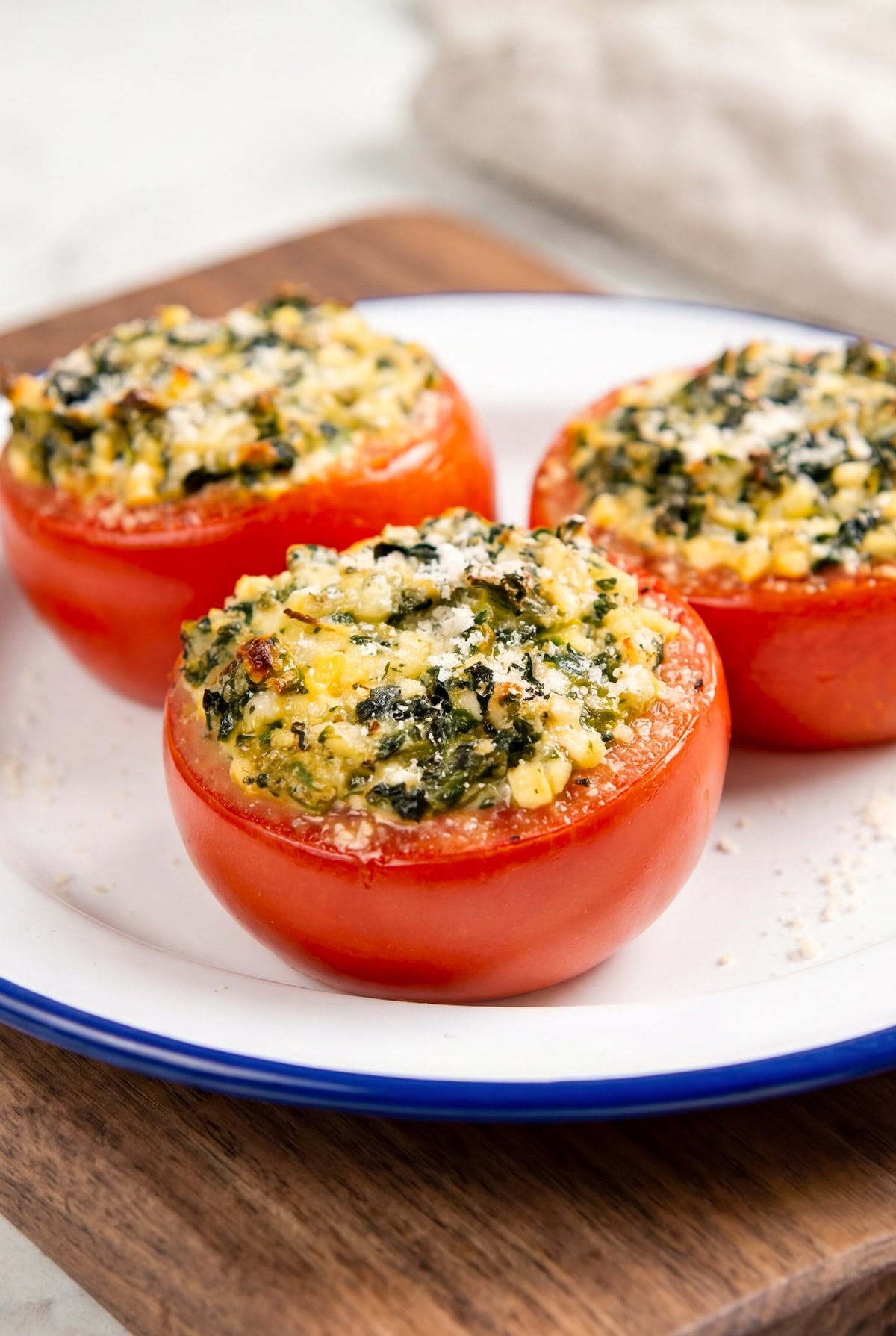 These stuffed tomatoes are ready in 30 minutes with a creamy spinach and cheese filling. Low carb, high protein, and seriously delicious! Three stuffed tomatoes with a creamy filling of spinach, cheese, and herbs are served on a white plate with a blue rim.