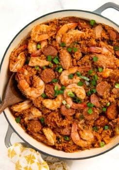 A pot of authentic cajun jambalaya filled with cooked shrimp, sliced sausage, and rice, garnished with chopped green onions. A wooden spoon rests in the pot, and a patterned cloth is nearby.