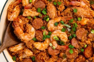A pot of authentic cajun jambalaya filled with cooked shrimp, sliced sausage, and rice, garnished with chopped green onions. A wooden spoon rests in the pot, and a patterned cloth is nearby.