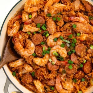 A pot of authentic cajun jambalaya filled with cooked shrimp, sliced sausage, and rice, garnished with chopped green onions. A wooden spoon rests in the pot, and a patterned cloth is nearby.