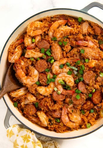 A pot of authentic cajun jambalaya filled with cooked shrimp, sliced sausage, and rice, garnished with chopped green onions. A wooden spoon rests in the pot, and a patterned cloth is nearby.