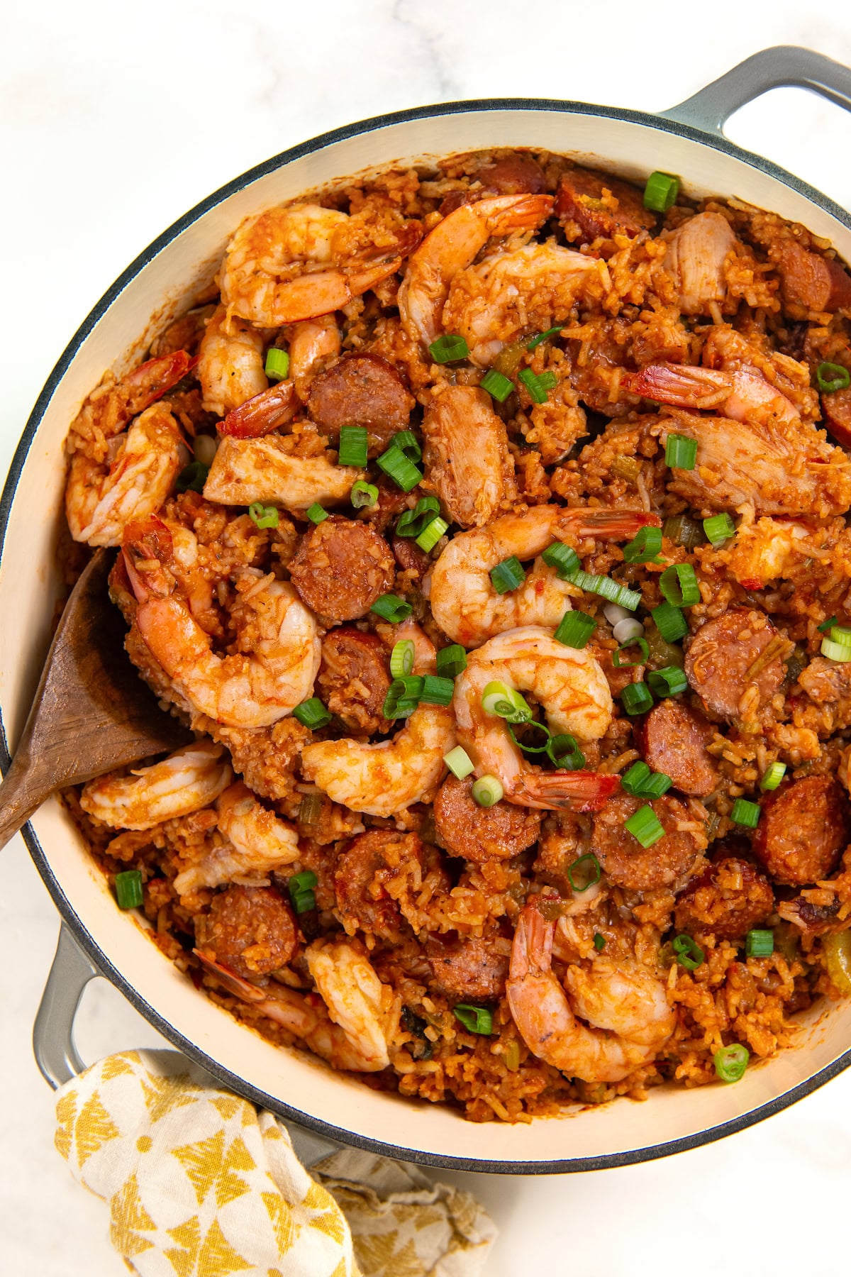 Full of bold Cajun flavor, this easy jambalaya recipe is a one-pot Louisiana classic loaded with chicken, andouille sausage, and shrimp. A pot of authentic cajun jambalaya filled with cooked shrimp, sliced sausage, and rice, garnished with chopped green onions. A wooden spoon rests in the pot, and a patterned cloth is nearby.