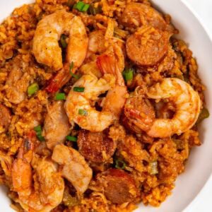 A white bowl filled with cajun jambalaya, featuring shrimp, sausage slices, diced vegetables, and seasoned rice in a reddish sauce, garnished with chopped green onions.