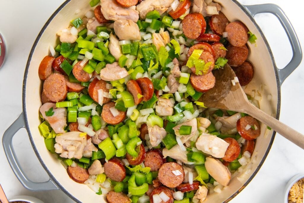 Full of bold Cajun flavor, this easy jambalaya recipe is a one-pot Louisiana classic loaded with chicken, andouille sausage, and shrimp. A large pot filled with sliced sausage, chopped chicken, green bell peppers, celery, and onions being stirred with a wooden spoon, showing the ingredients before cooking.