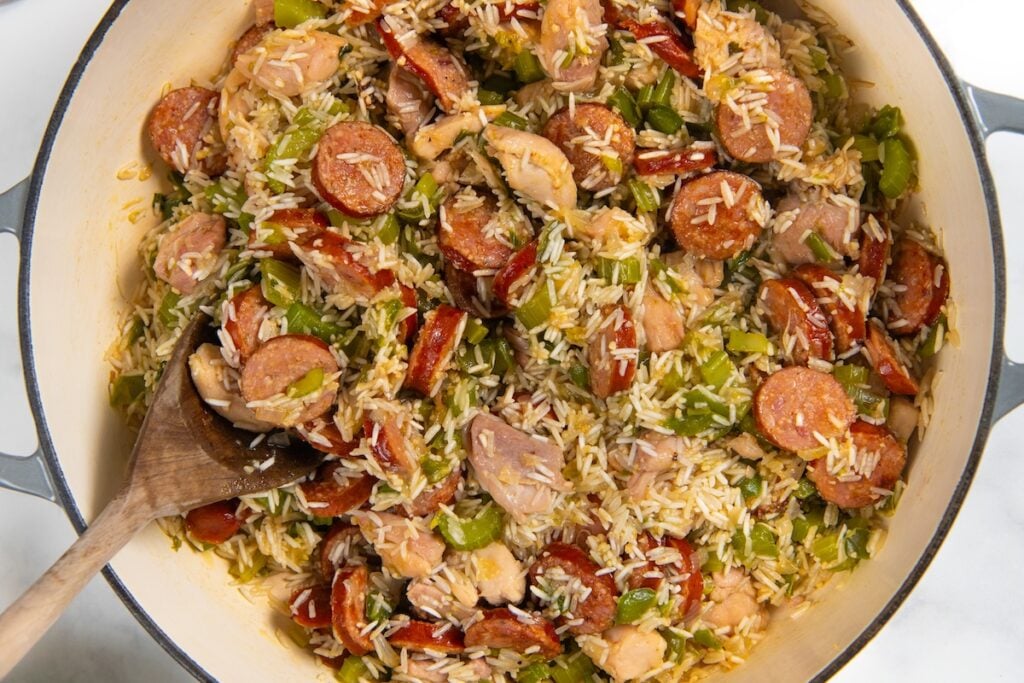 Full of bold Cajun flavor, this easy jambalaya recipe is a one-pot Louisiana classic loaded with chicken, andouille sausage, and shrimp. A pot filled with a mixture of sliced sausage, chicken pieces, rice, and chopped green bell peppers, with a wooden spoon resting inside.