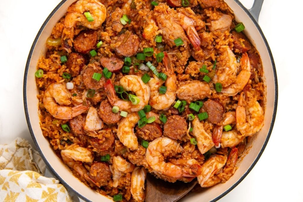 Full of bold Cajun flavor, this easy jambalaya recipe is a one-pot Louisiana classic loaded with chicken, andouille sausage, and shrimp. A skillet filled with cajun jambalaya, featuring shrimp, sliced sausage, rice, and vegetables, garnished with chopped green onions. A wooden spoon is partially visible in the dish.