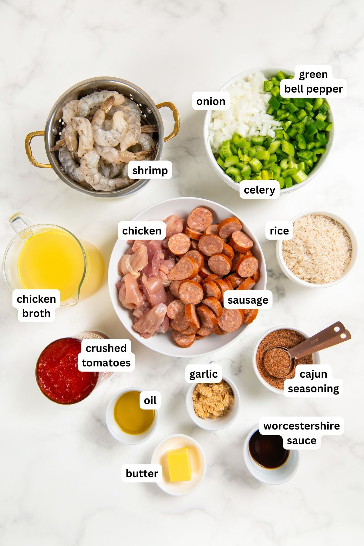 Full of bold Cajun flavor, this easy jambalaya recipe is a one-pot Louisiana classic loaded with chicken, andouille sausage, and shrimp. Top view of various labeled jambalaya recipe ingredients on a white surface, including shrimp, diced chicken, sausage, chopped onion, green bell pepper, celery, rice, chicken broth, crushed tomatoes, oil, garlic, butter, Cajun seasoning, and Worcestershire sauce.