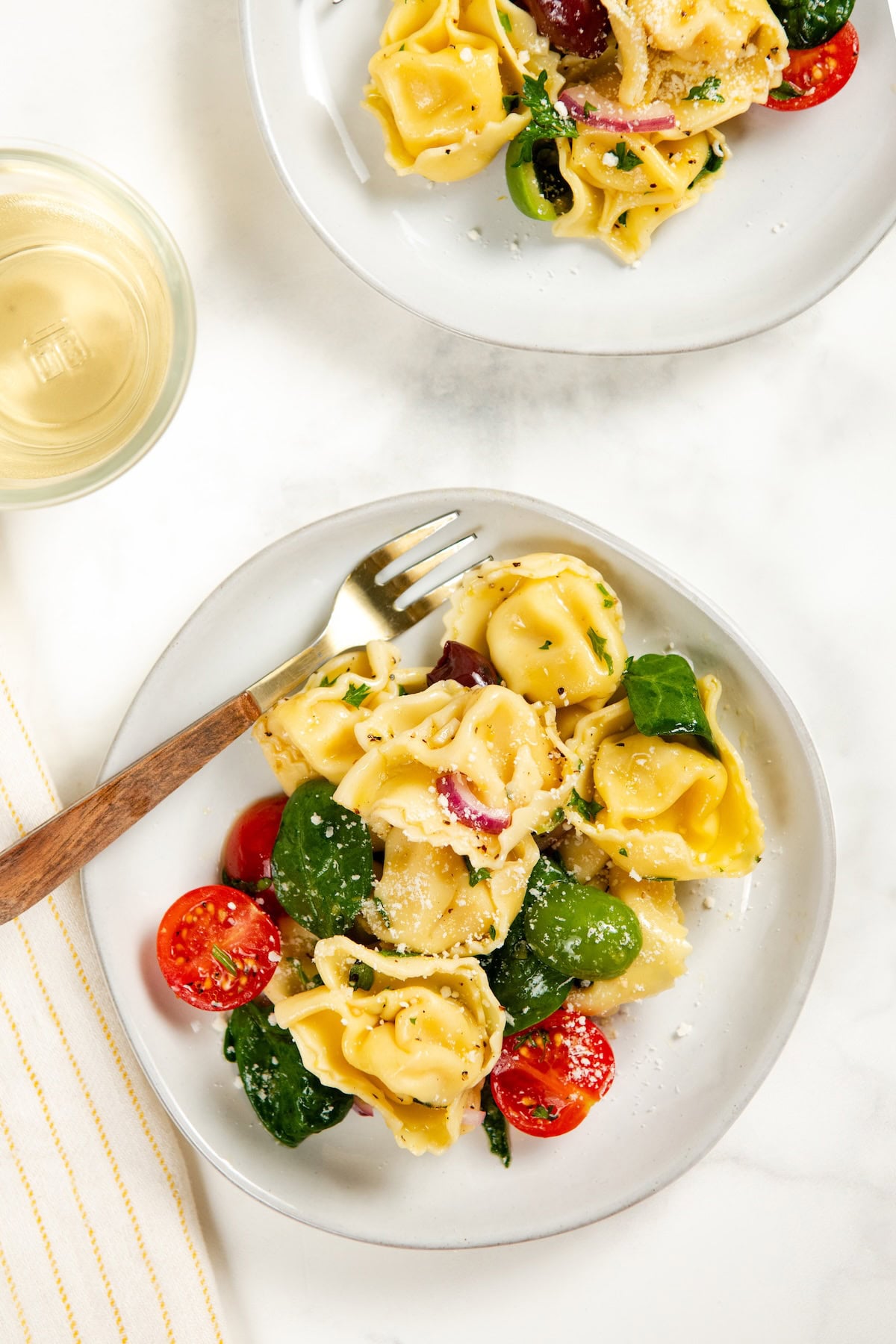 Cheese Tortellini Pasta Salad recipe with cherry tomatoes, spinach, olives, and a fresh lemon dressing is a summer favorite ready in just 15 minutes! A plate of tortellini pasta salad with spinach, cherry tomatoes, red onions, and olives, garnished with grated cheese. A fork rests on the plate, and a glass of white wine sits nearby on a white surface.