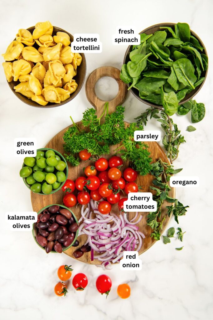 Cheese Tortellini Pasta Salad recipe with cherry tomatoes, spinach, olives, and a fresh lemon dressing is a summer favorite ready in just 15 minutes! A wooden board displays labeled tortellini pasta salad recipe ingredients: cheese tortellini, fresh spinach, parsley, oregano, cherry tomatoes, red onion slices, green olives, and kalamata olives.