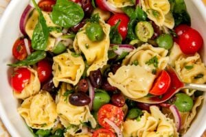 A white bowl filled with cheese tortellini pasta salad, featuring cherry tomatoes, spinach, sliced red onions, green and black olives, and fresh herbs on a patterned tablecloth.