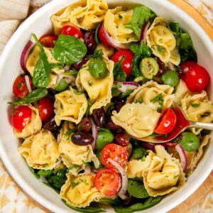 A white bowl filled with cheese tortellini pasta salad, featuring cherry tomatoes, spinach, sliced red onions, green and black olives, and fresh herbs on a patterned tablecloth.