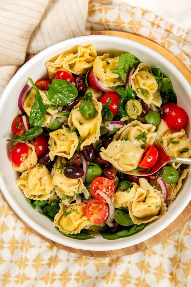 A white bowl filled with cheese tortellini pasta salad, featuring cherry tomatoes, spinach, sliced red onions, green and black olives, and fresh herbs on a patterned tablecloth.