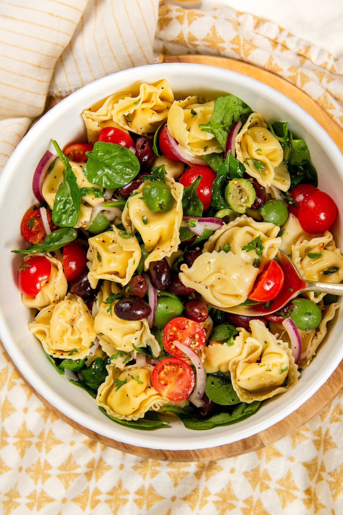 Cheese Tortellini Pasta Salad recipe with cherry tomatoes, spinach, olives, and a fresh lemon dressing is a summer favorite ready in just 15 minutes! A white bowl filled with cheese tortellini pasta salad, featuring cherry tomatoes, spinach, sliced red onions, green and black olives, and fresh herbs on a patterned tablecloth.