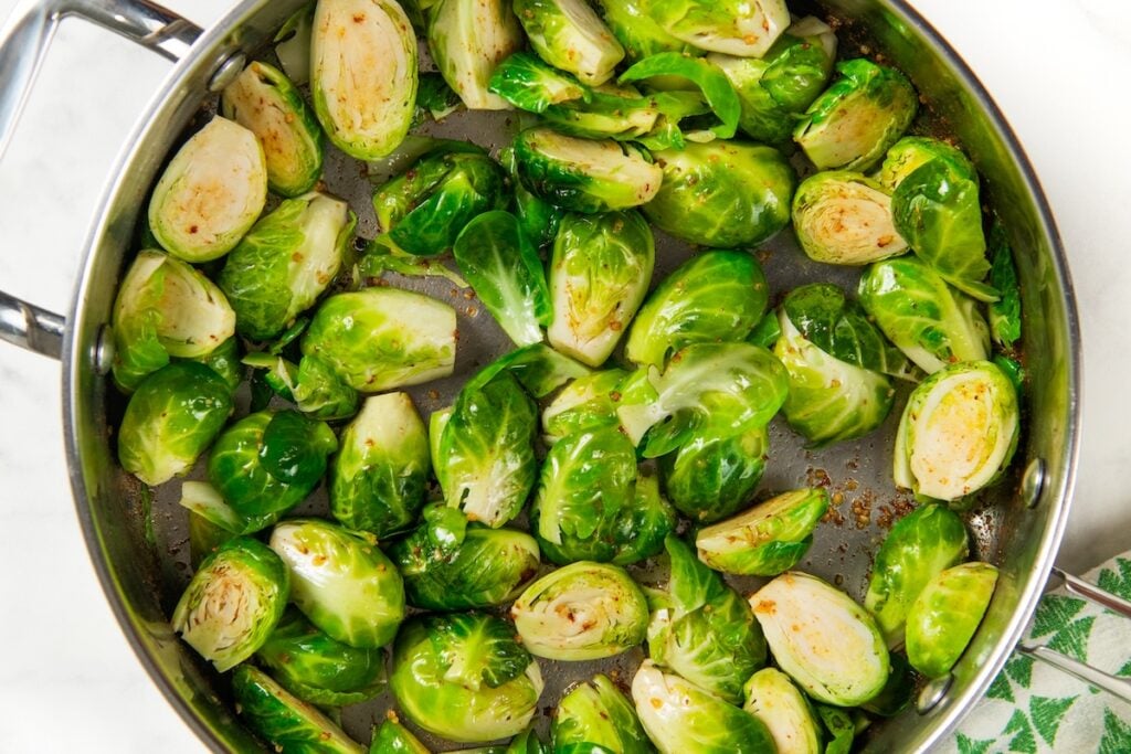 Hot Honey Brussels Sprouts with crispy bacon, caramelized sprouts, and a sweet-spicy honey drizzle. One skillet, ready in 25 minutes! Brussels sprouts cut in half and sautéed in a large stainless steel skillet with garlic and seasonings, showing slight browning on some pieces.