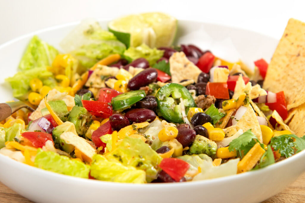 This hearty Ground Beef Taco Salad recipe combines all the best taco flavors in one bowl with a fresh cilantro lime dressing! Ready in 30 minutes. A colorful taco salad with lettuce, kidney beans, grilled chicken, corn, jalapeño slices, diced tomatoes, shredded cheese, cilantro, tortilla chips, and a lime wedge in a white bowl.