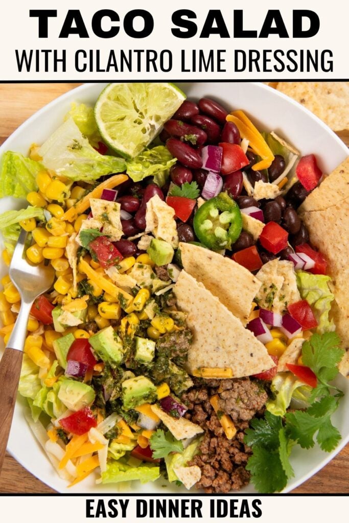 A colorful taco salad in a bowl with ground beef, lettuce, black beans, corn, tomatoes, jalapeños, cilantro, tortilla chips, and a lime wedge, topped with cilantro lime dressing. Text reads Taco Salad with Cilantro Lime Dressing.