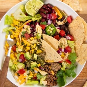 A taco salad in a white bowl with ground beef, lettuce, black beans, corn, tomatoes, avocado, shredded cheese, red onion, jalapeño, tortilla chips, cilantro, and a lime wedge on top. A fork and extra chips are nearby.