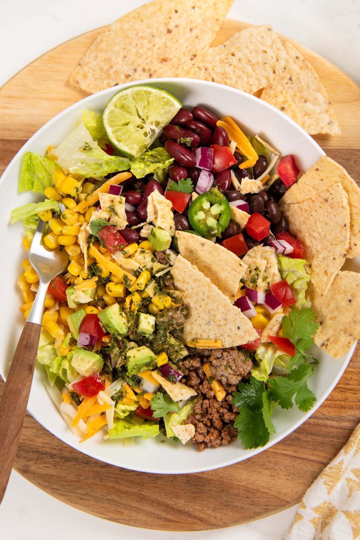 This hearty Ground Beef Taco Salad recipe combines all the best taco flavors in one bowl with a fresh cilantro lime dressing! Ready in 30 minutes. A taco salad in a white bowl with ground beef, lettuce, black beans, corn, tomatoes, avocado, shredded cheese, red onion, jalapeño, tortilla chips, cilantro, and a lime wedge on top. A fork and extra chips are nearby.