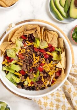 A ground beef taco salad in a white bowl with ground beef, lettuce, corn, black beans, kidney beans, shredded cheese, diced tomatoes, and tortilla chips arranged around the edge. Sliced avocado and lime are on the side.
