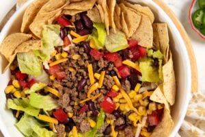 A ground beef taco salad in a white bowl with ground beef, lettuce, corn, black beans, kidney beans, shredded cheese, diced tomatoes, and tortilla chips arranged around the edge. Sliced avocado and lime are on the side.
