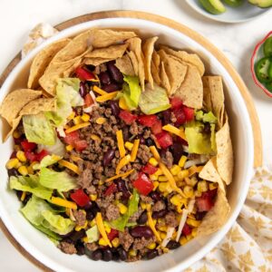 A ground beef taco salad in a white bowl with ground beef, lettuce, corn, black beans, kidney beans, shredded cheese, diced tomatoes, and tortilla chips arranged around the edge. Sliced avocado and lime are on the side.