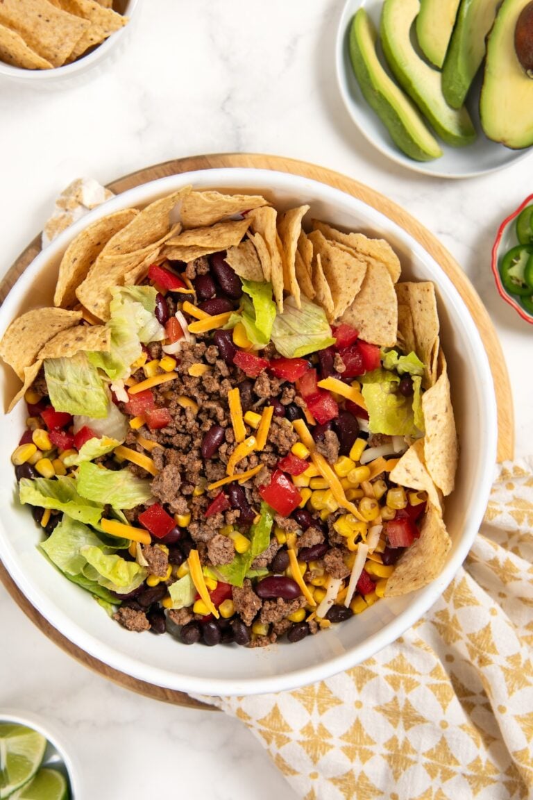 A ground beef taco salad in a white bowl with ground beef, lettuce, corn, black beans, kidney beans, shredded cheese, diced tomatoes, and tortilla chips arranged around the edge. Sliced avocado and lime are on the side.