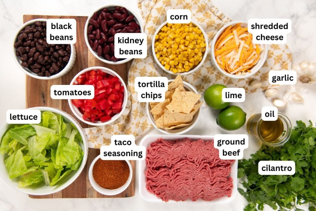 This hearty Ground Beef Taco Salad recipe combines all the best taco flavors in one bowl with a fresh cilantro lime dressing! Ready in 30 minutes. An overhead view of taco salad recipe ingredients in bowls on a white surface, including black beans, kidney beans, corn, shredded cheese, tomatoes, garlic, oil, lettuce, taco seasoning, tortilla chips, lime, ground beef, and cilantro, each labeled.
