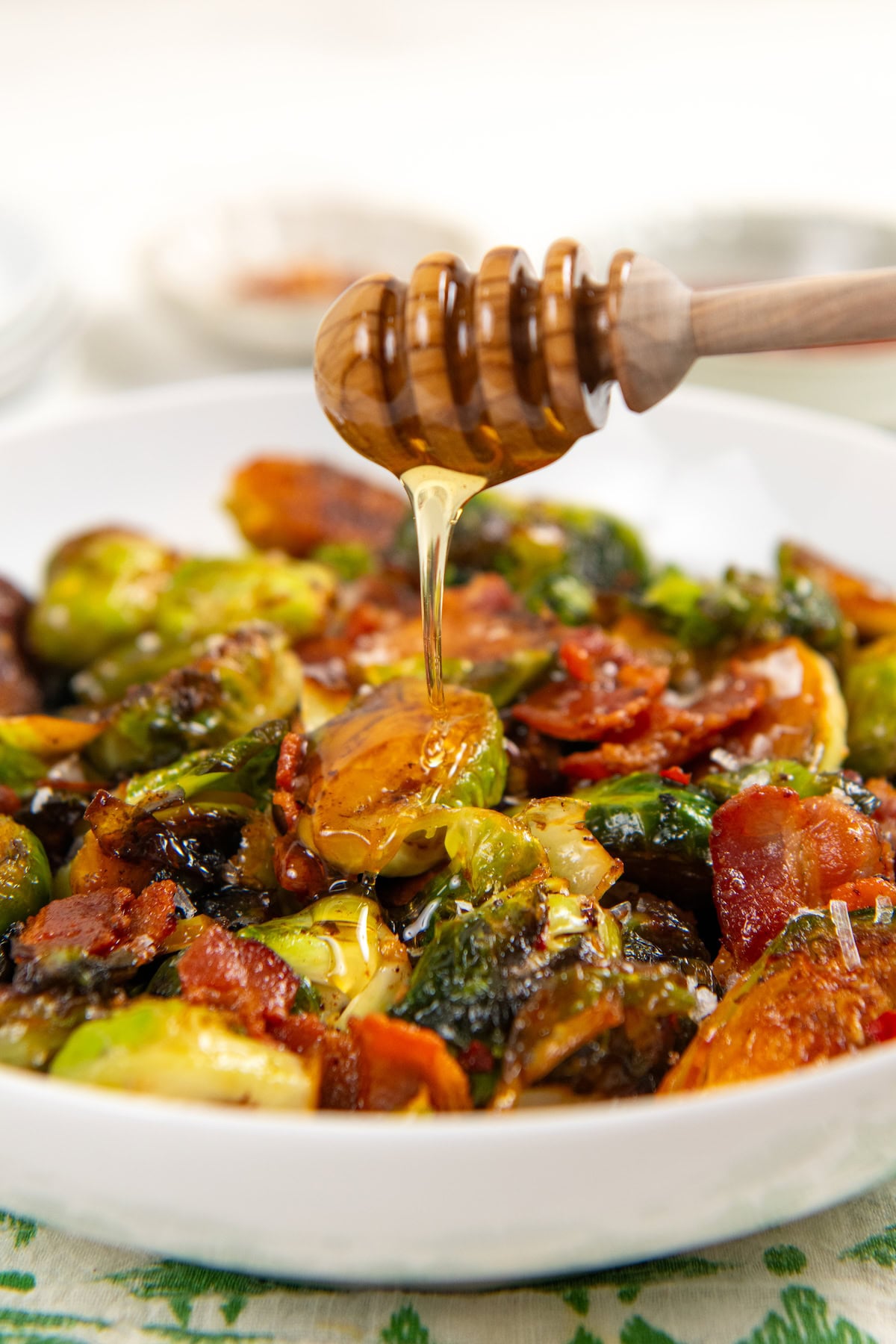 Hot Honey Brussels Sprouts with crispy bacon, caramelized sprouts, and a sweet-spicy honey drizzle. One skillet, ready in 25 minutes! A honey dipper drizzles hot honey over a bowl of roasted Brussels sprouts with crispy bacon.